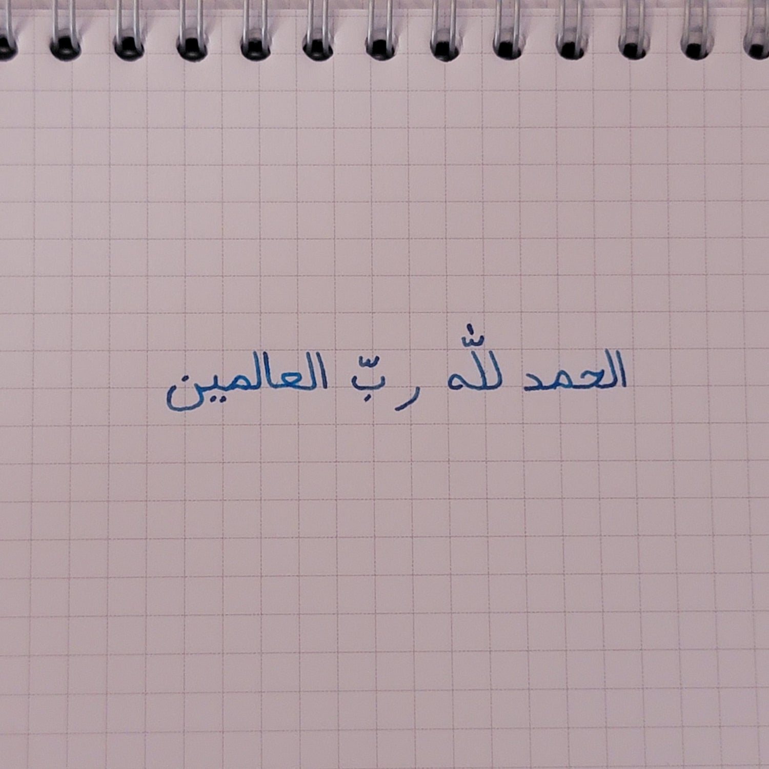 Photographie d'une feuille de carnet spirale à petits carreaux, où il est écrit en arabe : الحمد للّٰه ربّ العالمينن