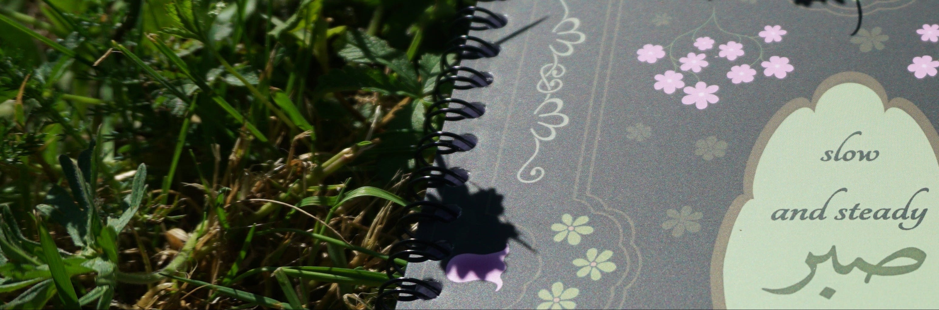 Photographie d'un carnet illustré de fleurs et d'inscriptions en arabe et en anglais, dans l'herbe, à la lumière du jour.