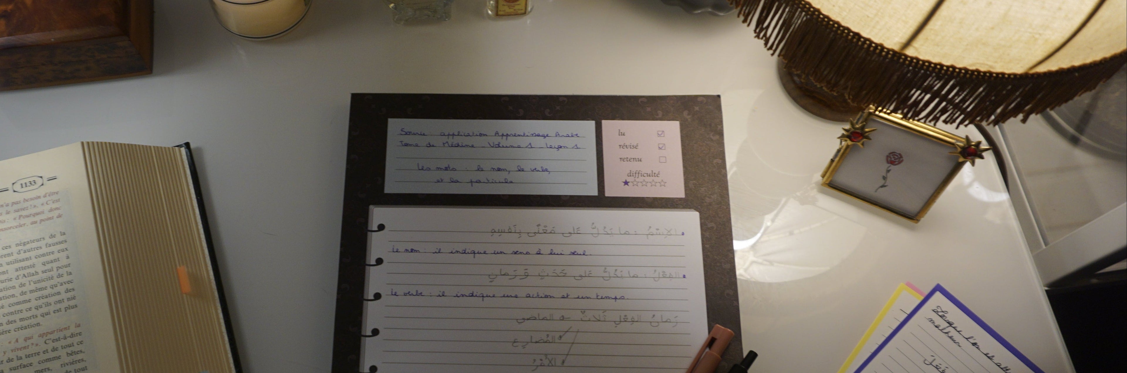 Photographie d'un bloc-notes de révisions dont la page a été remplie à la main par une leçon de grammaire arabe. Le bloc-notes est sur un bureau entouré d'autres objets de bureau et de décoration.